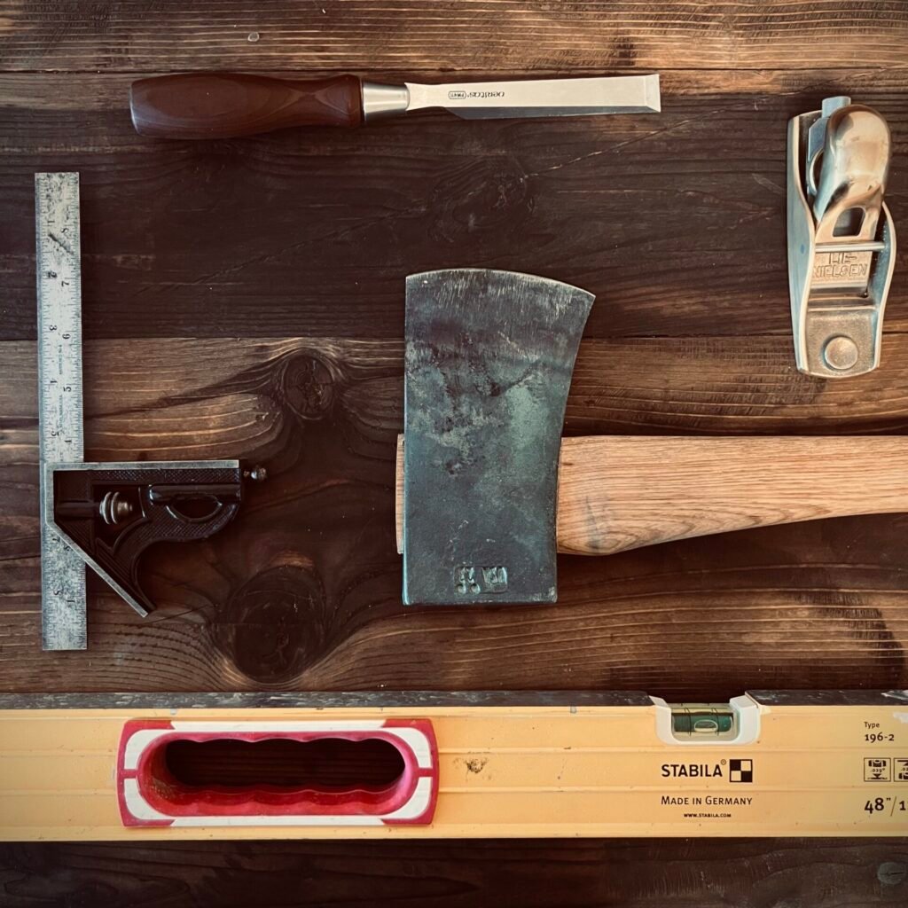 Heirloom-quality hand tools including a forged axe, wood chisel, hand plane, square, and Stabila level arranged on a wooden workbench